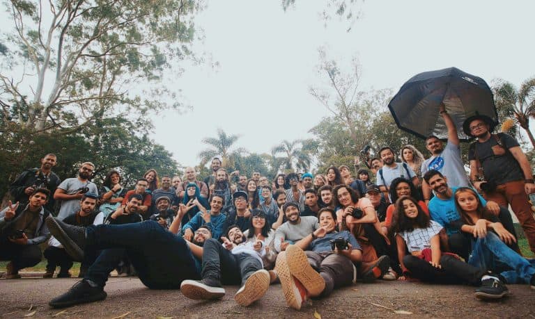 People group photo outdoors with lush trees, bright sky, diverse individuals, some holding cameras, capturing a joyful moment during a sunny day.